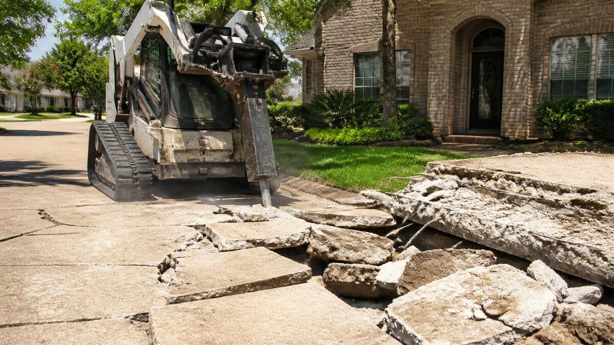 A skid steer with a breaker breaking up a cracked concrete driveway in front of a brick house