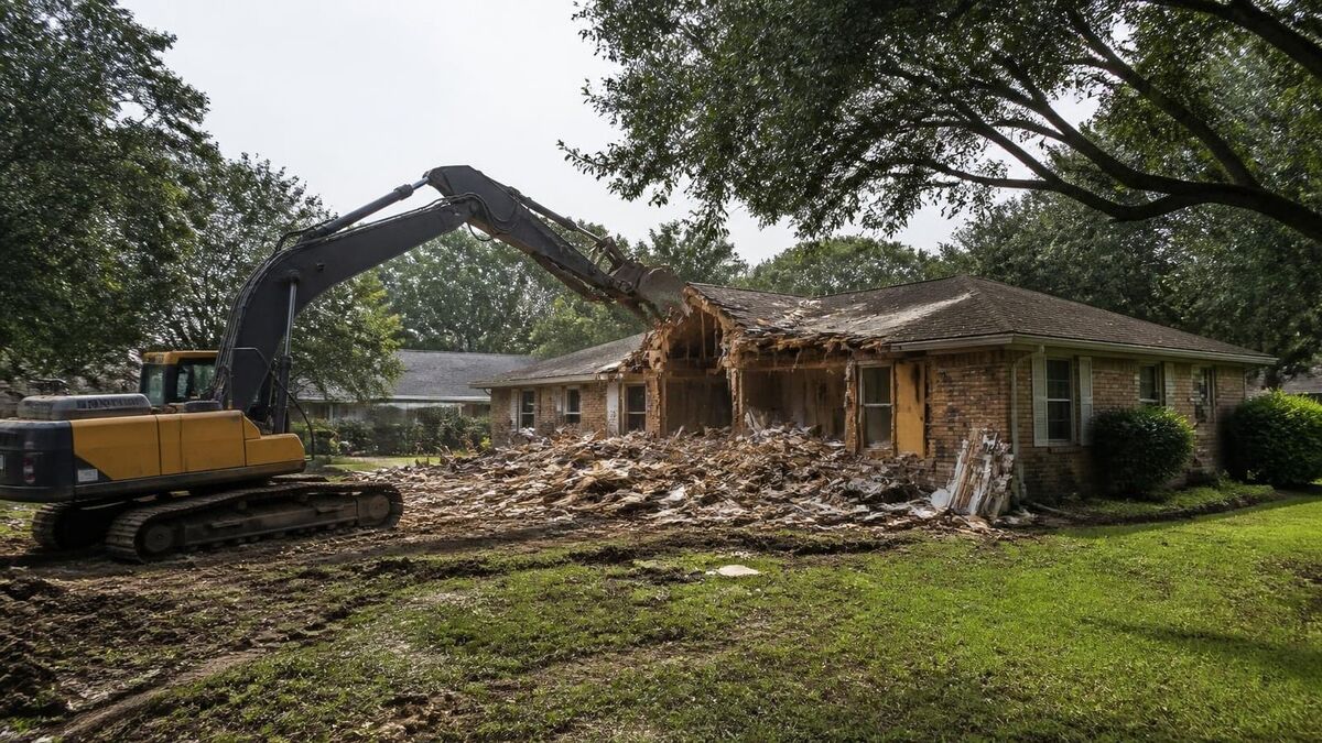 An excavator tearing down an old brick ranch house on a flat suburban lot, with a pile of debris and oak trees around it